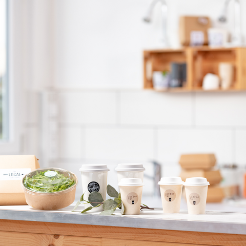 Een houten tafel met een kom salade, verschillende koffiekopjes en een verpakking, omringd door een lichte, moderne keuken.