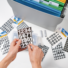 Handen houden een zwart bord met witte cijfers. Op de achtergrond zijn er meer cijfers en letters te zien, evenals een opbergsysteem.