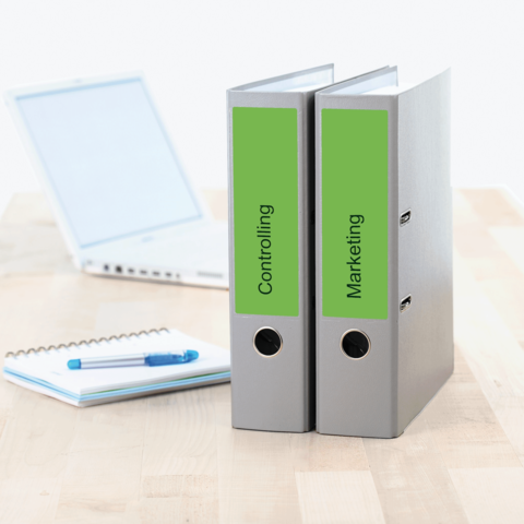 Zwei Aktenordner mit den Beschriftungen "Controlling" und "Marketing" stehen neben einem Laptop und einem Notizblock auf einem Tisch.