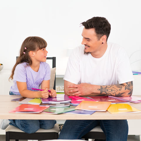 Ein Mann und ein Mädchen sitzen an einem Tisch, umgeben von bunten Farbmusterkarten, während sie miteinander sprechen.