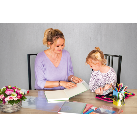 Eine Frau und ein Mädchen sitzen an einem Tisch und betrachten gemeinsam ein Buch. Auf dem Tisch liegen verschiedene Schreibmaterialien und Blumen.