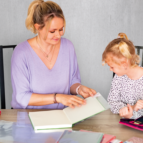 Eine Frau und ein Kind sitzen an einem Tisch und schauen gemeinsam in ein Buch. Auf dem Tisch liegen weitere Bücher und Bastelmaterialien.