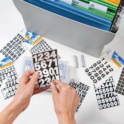 Handen houden een zwart bord met witte cijfers, omringd door verschillende genummerde en alfabetische stickers in een organizer.
