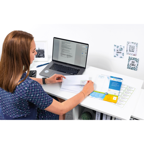 Eine Person sitzt an einem Schreibtisch und arbeitet am Laptop, während sie Dokumente und Briefe sortiert. Auf dem Tisch liegen verschiedene Unterlagen.