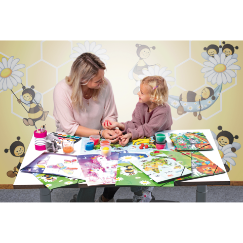 Eine Frau und ein Kind sitzen an einem Tisch, umgeben von Bastelmaterialien und Kunstwerken. Sie interagieren freundlich miteinander.