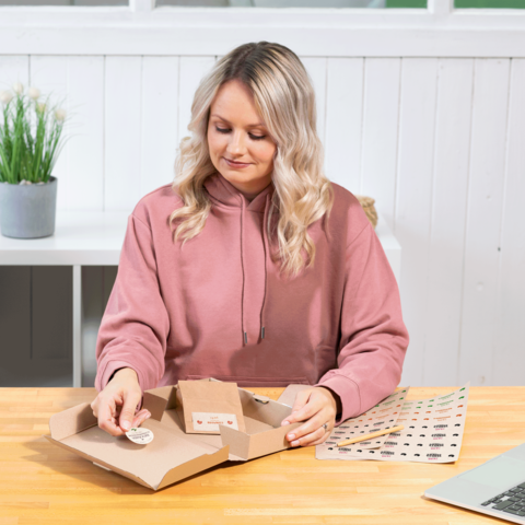 Eine Frau mit lockigem Haar in einem rosa Hoodie öffnet eine Pappverpackung und betrachtet deren Inhalt auf einem Tisch.