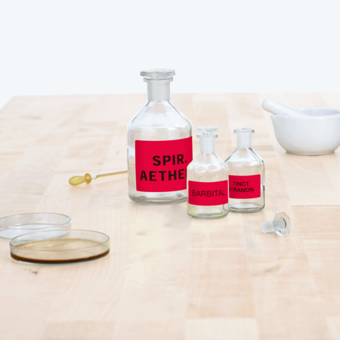 Flaschen mit roten Etiketten, beschriftet mit "Spir. Aether", "Barbital" und "Tinct. Tramon", auf einem Holzuntergrund.