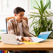 Ein junger Mann sitzt an einem Schreibtisch, arbeitet mit einem Laptop und blättert durch Dokumente in einer Ordner. Pflanzen sind im Hintergrund sichtbar.