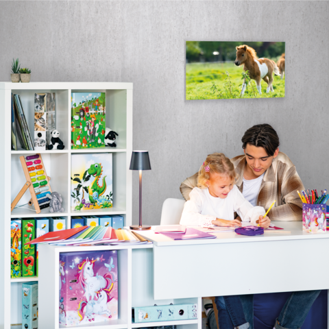 Ein Kind und ein Jugendlicher sitzen an einem Schreibtisch und malen. Im Hintergrund steht ein Regal mit Spielzeug und Bastelmaterial.