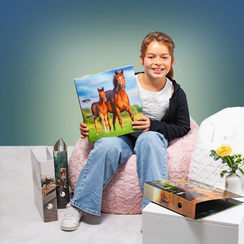 Een meisje zit op een stoel en houdt een foto van twee paarden in haar hand. Naast haar staan mappen en een plant.