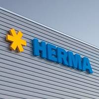 Geel sterrenlogo's en blauwe letters vormen het woord 'HERMA' op de buitenmuur van een modern gebouw.