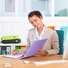 Eine Frau in einem hellen Anzug sitzt an einem Schreibtisch und betrachtet einen Ordner, während sie Notizen macht.