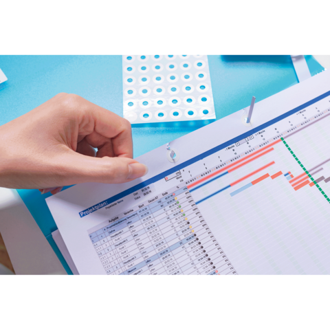 Eine Hand hält ein Dokument mit einem Zeitplan, das auf einem Tisch liegt. Im Hintergrund sind bunte Aufkleber sichtbar.