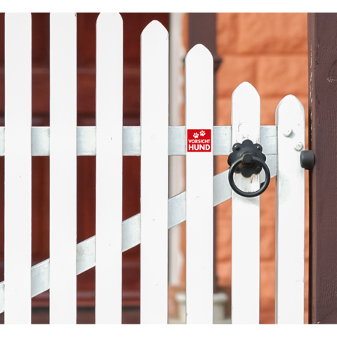 Weißer Gartenzaun mit einem roten Schild, das auf einen Hund hinweist. Der Zaun hat einen runden Türknauf.