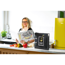 Eine Frau mit Brille steht in einer modernen Küche und hält ein Kochbuch. Auf der Arbeitsfläche liegen frisches Gemüse und Tomaten.