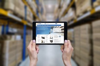 Tablet in de hand, toont een website met informatie over voorraadbeheer, op de achtergrond rekken met dozen.