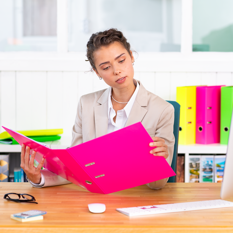 Eine Frau in einem Büro betrachtet einen pinken Ordner, während sie an einem Schreibtisch sitzt. Im Hintergrund sind bunte Aktenordner sichtbar.