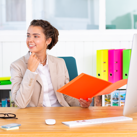 Eine Frau in einem Büro sitzt am Schreibtisch, hält einen orangefarbenen Ordner und lächelt, während sie nachdenklich aussieht.