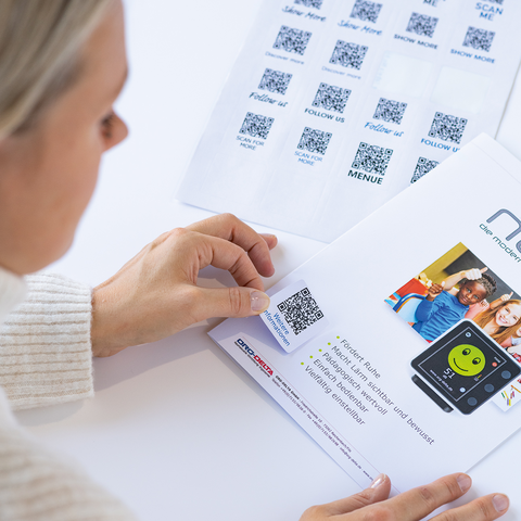 Eine Person scannt einen QR-Code auf einem Menüblatt, während ein weiteres Blatt mit mehreren QR-Codes im Hintergrund liegt.