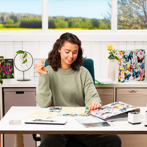 Een jonge vrouw zit aan een tafel, omringd door notities en een laptop, terwijl ze met één hand een gebaar maakt. Op de achtergrond zijn er ramen met uitzicht op een groene landschap.