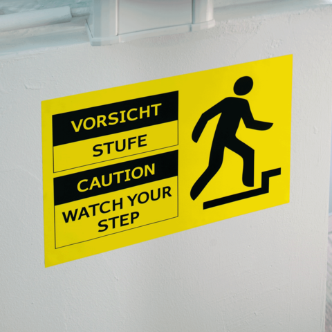 Warnschild mit den Hinweisen "Vorsicht Stufe", "Caution" und "Watch Your Step", das auf eine Stufe hinweist.