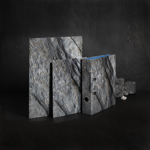 Drei verschiedene Platten aus grauem Gestein, eine mit blauer Kante, stehen auf einer dunklen Oberfläche. Im Hintergrund sind kleine Gesteinsstücke sichtbar.