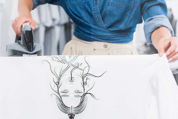 Een persoon strijkt een wit shirt met een grafische print terwijl hij op een strijkplank staat.