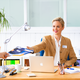 Een vrouw in een kantoor geeft een man een document, terwijl ze aan een bureau zit met een laptop en kantoormateriaal.