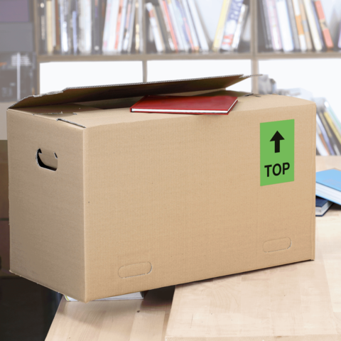 Kartonbox auf einem Tisch, mit einem roten Heftchen oben und einem grünen "TOP"-Schild. Im Hintergrund sind Bücherregale sichtbar.