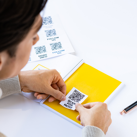 Eine Person klebt einen QR-Code auf eine gelbe Seite eines Notizbuchs, während sie einen Stift in der Hand hält.