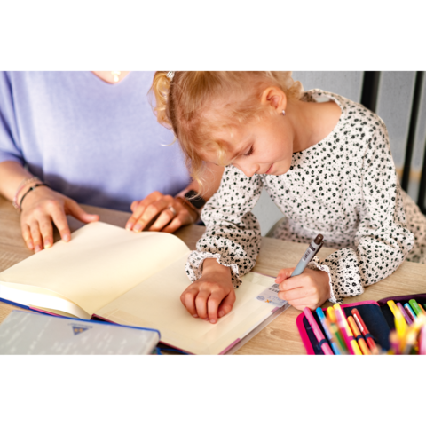 Ein kleines Mädchen schreibt in ein Notizbuch, während eine erwachsene Hand daneben hilft. Bunte Stifte liegen auf dem Tisch.
