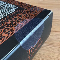 Oplossingen Cosmetica: Zwarte verpakking met decoratieve, oranje patronen en tekst. De flap van de verpakking is verzegeld met een transparante sluitsticker.