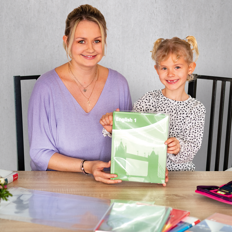 Eine Frau und ein Mädchen sitzen an einem Tisch und halten ein grünes Heft. Auf dem Tisch liegen Schreibmaterialien und Blumen.