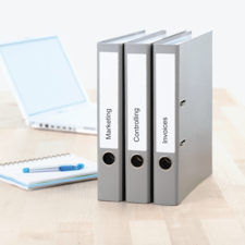 Drei graue Ordner mit Beschriftungen "Marketing", "Controlling" und "Rechnungen" stehen auf einem Tisch neben einem Laptop und einem Notizblock.