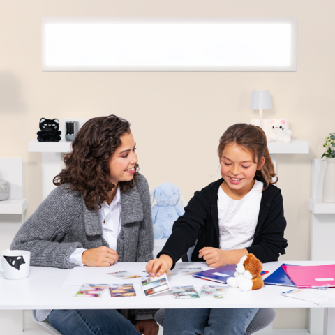 Twee meisjes zitten aan een tafel, omringd door foto's en notitiemateriaal, terwijl ze met elkaar praten en lachen.