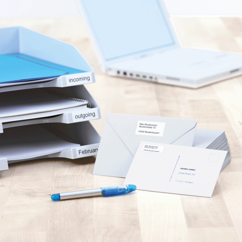 Ordner mit beschrifteten Fächern für eingehende und ausgehende Dokumente, daneben ein Stift und mehrere Umschläge mit Adressetiketten.