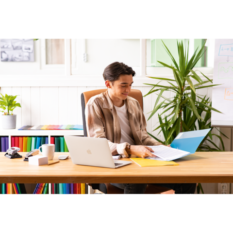 Ein junger Mann sitzt an einem Schreibtisch, arbeitet mit einem Laptop und blättert durch Dokumente in einer Ordnermappe. Pflanzen sind im Hintergrund.