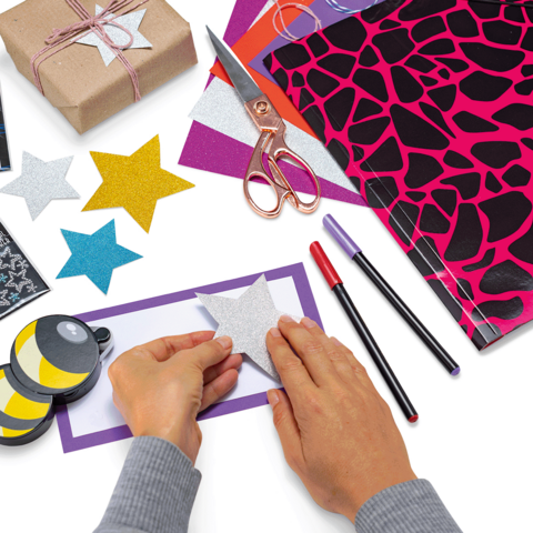 Ein Schreibtisch mit bunten Ordnern, Schreibmaterialien, einer Lampe und einer kleinen Pflanze. Im Hintergrund sind Fenster mit Tageslicht sichtbar.