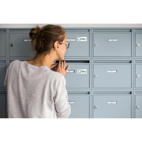 Eine Frau mit Brille schaut in einen grauen Briefkasten. Im Hintergrund sind mehrere ähnliche Briefkästen zu sehen.