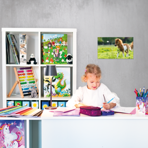 Ein Mädchen sitzt an einem Schreibtisch, schreibt in ein Heft und hat eine Federmappe vor sich. Im Hintergrund sind Regale mit Spielzeug und Kunstmaterial.