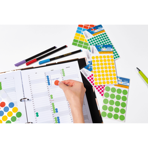 Ein Hand greift nach einem roten Aufkleber, während ein Planer mit farbigen Markierungen und mehreren Aufkleberbögen auf dem Tisch liegt.