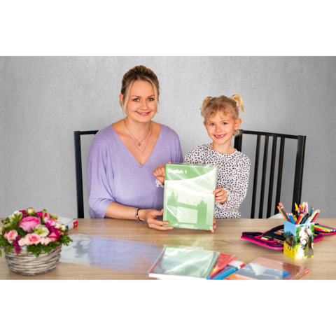 Eine Frau und ein Mädchen sitzen an einem Tisch, halten ein grünes Heft und lächeln. Auf dem Tisch liegen Schreibmaterialien und Blumen.