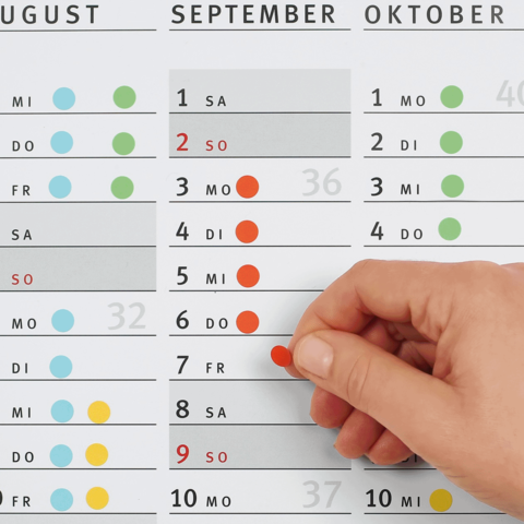 Ein Hand, die einen roten Punkt auf einem Kalender für September und Oktober platziert, mit hervorgehobenen Daten und Wochentagen.