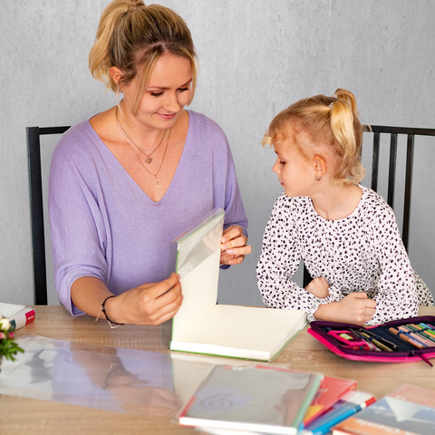 Eine Frau und ein kleines Mädchen sitzen an einem Tisch, während sie gemeinsam an einem Kunstprojekt arbeiten. Blumen und Bastelmaterial sind sichtbar.
