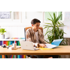 Ein junger Mann sitzt an einem Schreibtisch, arbeitet mit einem Laptop und blättert durch Dokumente in einer Ordner. Pflanzen und bunte Aktenordner sind im Hintergrund sichtbar.